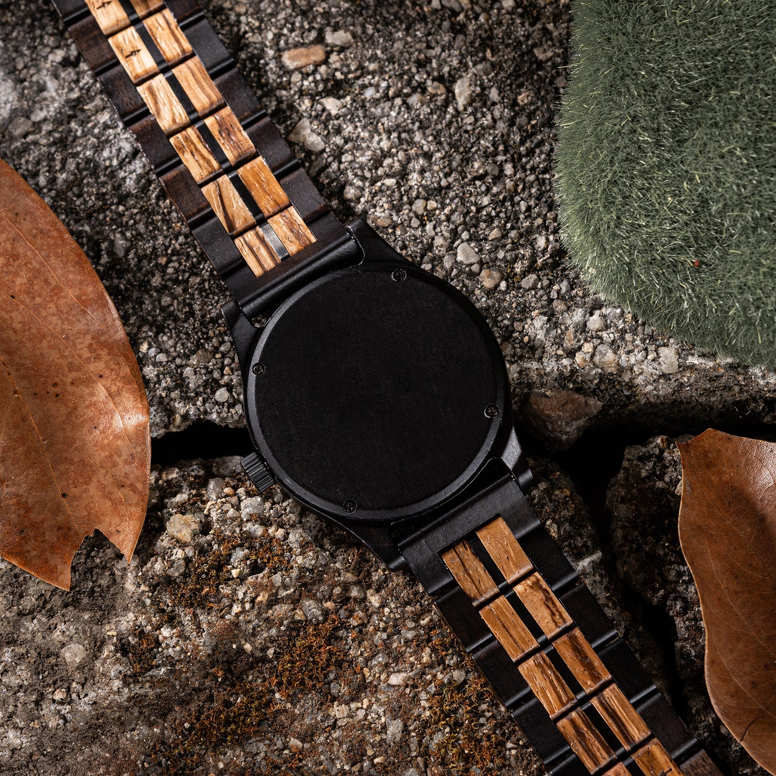 Eclipse Noir men's watch, black stainless steel case, minimalist dial, and two-tone dark and light wood strap, shown from the back view on a rocky surface.