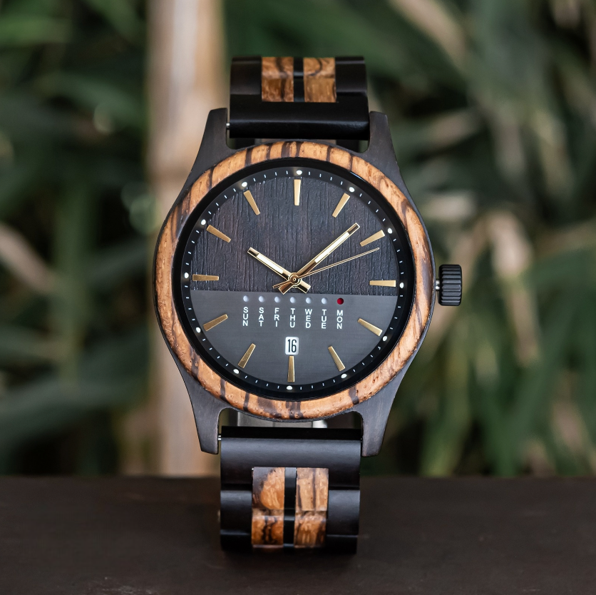Front view of a dark wooden watch with a black dial and gold markers, against a blurred background of bamboo stalks.