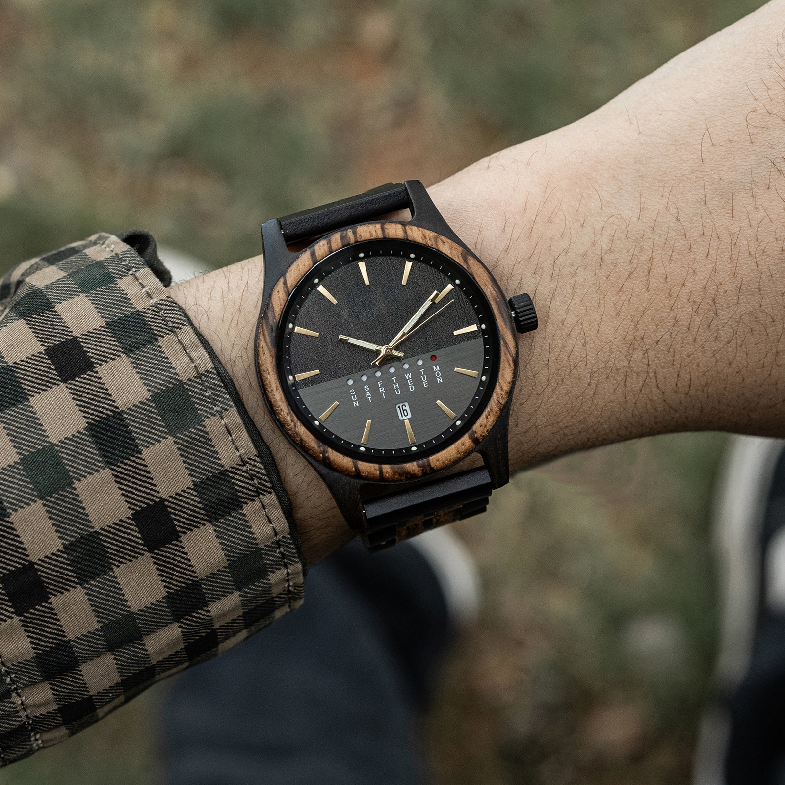 A man's wrist wearing a dark wooden watch with a black dial and gold markers. He is wearing a plaid shirt, with a blurred green outdoor background.