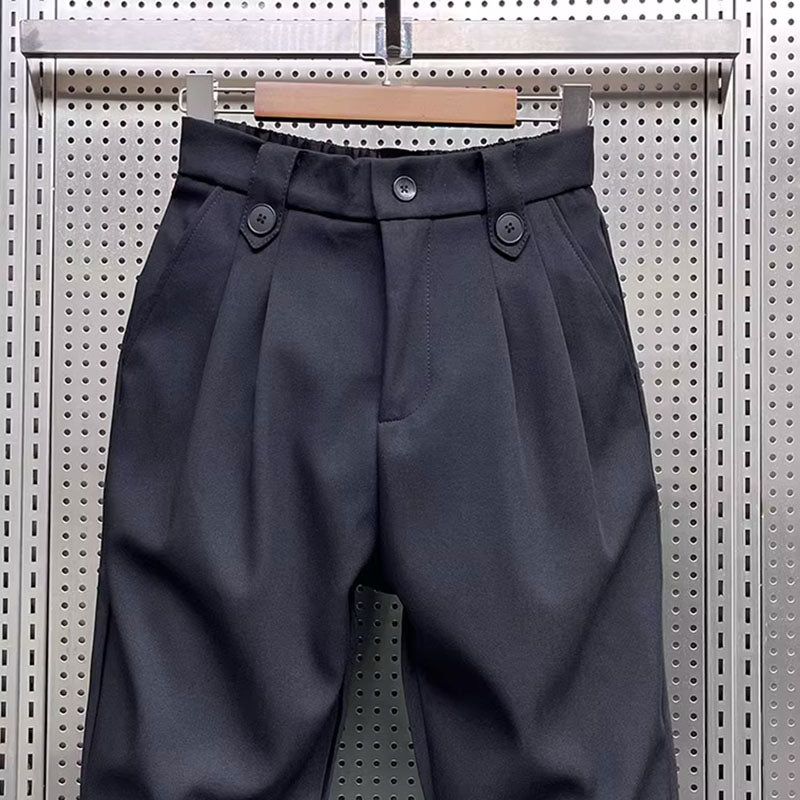 Close-up view of the waist of black pleated harem trousers, showing the button closure, belt loops, and fabric pleats.