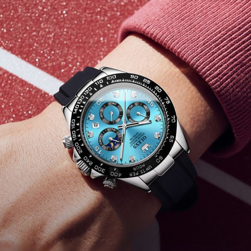 A man's wrist showcasing an Elysium Timepiece men's watch with a blue dial, silver case, and black silicone strap, against a textured red background.