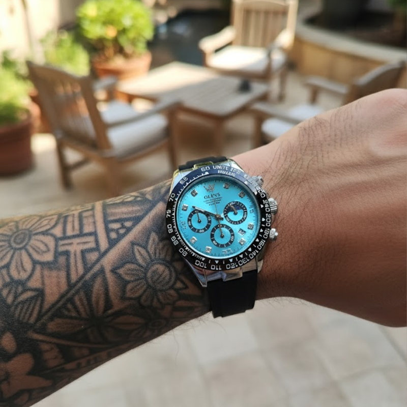 A close-up of a man's tattooed arm wearing an Elysium Timepiece men's watch with a blue dial, silver case, and black silicone strap, shown outdoors with blurred patio furniture in the background.