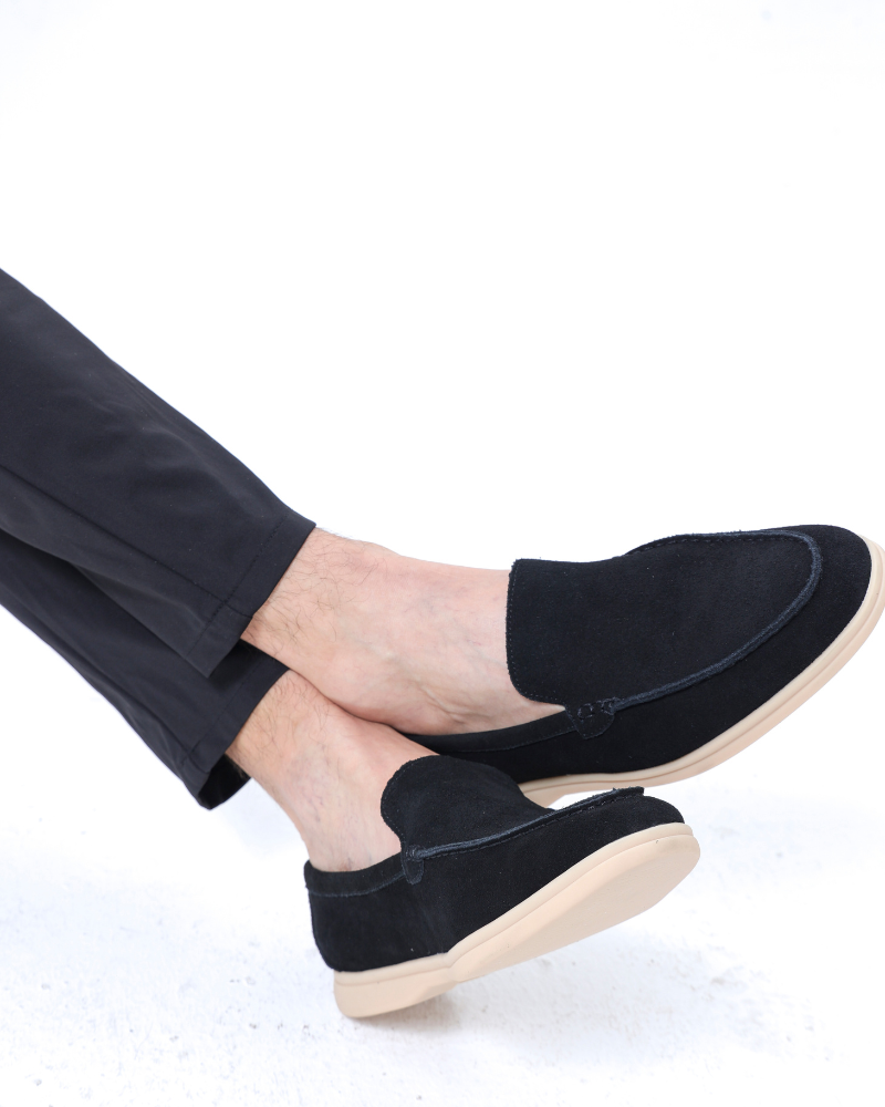 A man's crossed legs wearing Heritage Luxe black suede loafers with classic stitching and a tan sole, paired with black pants, on a clean white background.