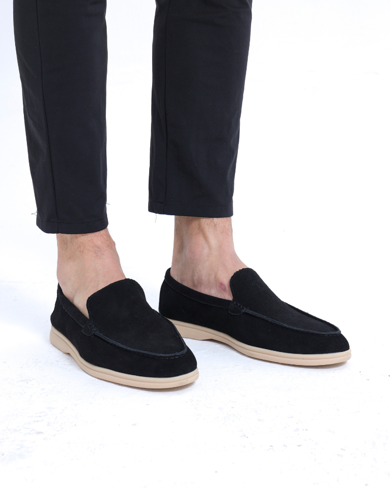 A man's feet wearing Heritage Luxe black suede loafers with classic stitching and a tan sole, paired with black pants, standing on a clean white background.
