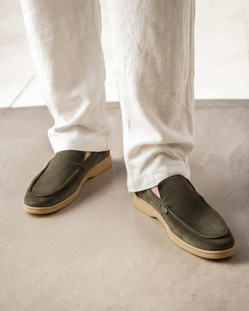 Men's Heritage Luxe olive green suede loafers with classic stitching, worn by a person in white linen trousers, standing on a light floor.
