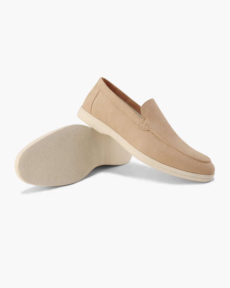 A pair of Heritage Luxe beige suede loafers, one upright showing the side profile and classic stitching, the other on its side revealing the textured sole pattern.