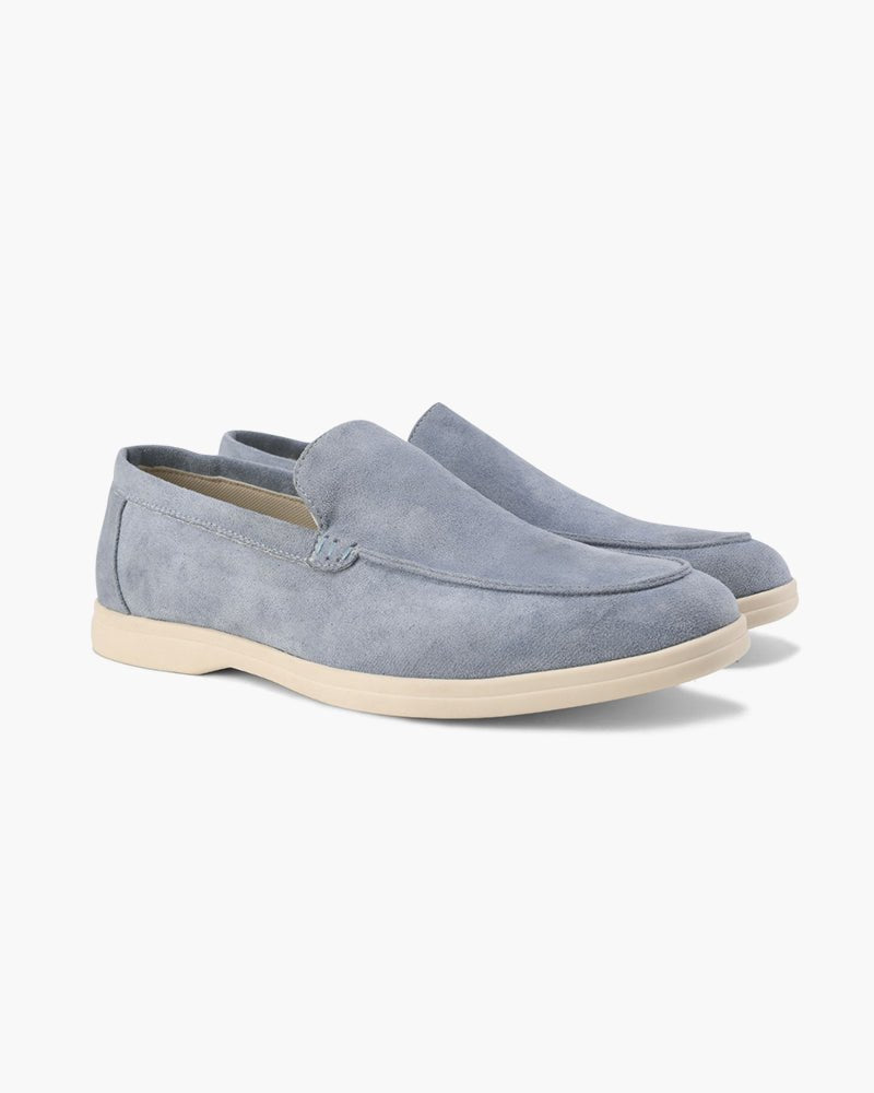 A pair of Heritage Luxe Fog Blue suede loafers with classic stitching, featuring a light cream sole, presented on a clean white background. These men's casual slip-ons are stylish and comfortable.