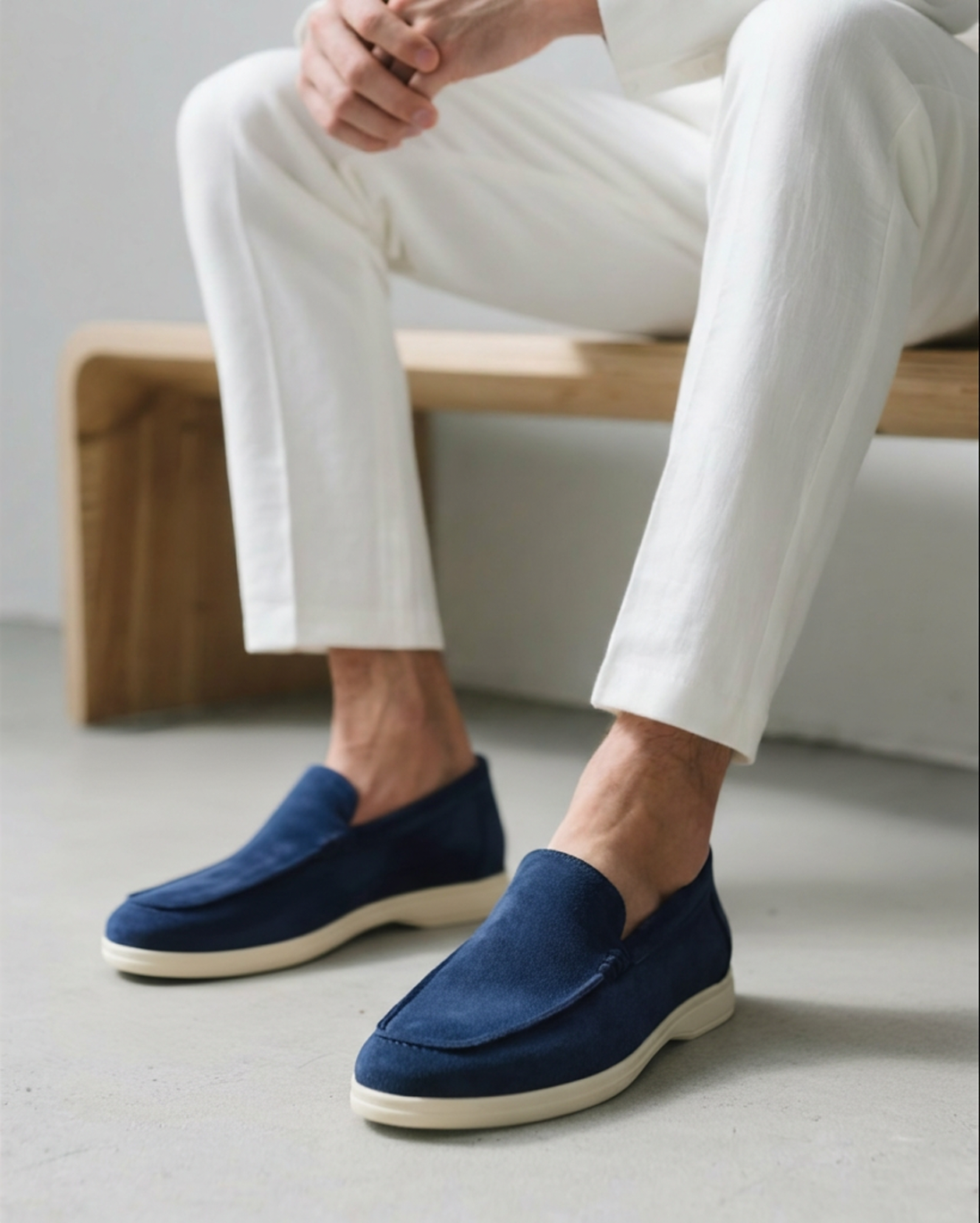 A man wearing Heritage Luxe Marine Blue suede loafers with cream soles and white linen pants, sitting casually on a light wooden bench. These comfortable men's luxury loafers are perfect everyday wear.