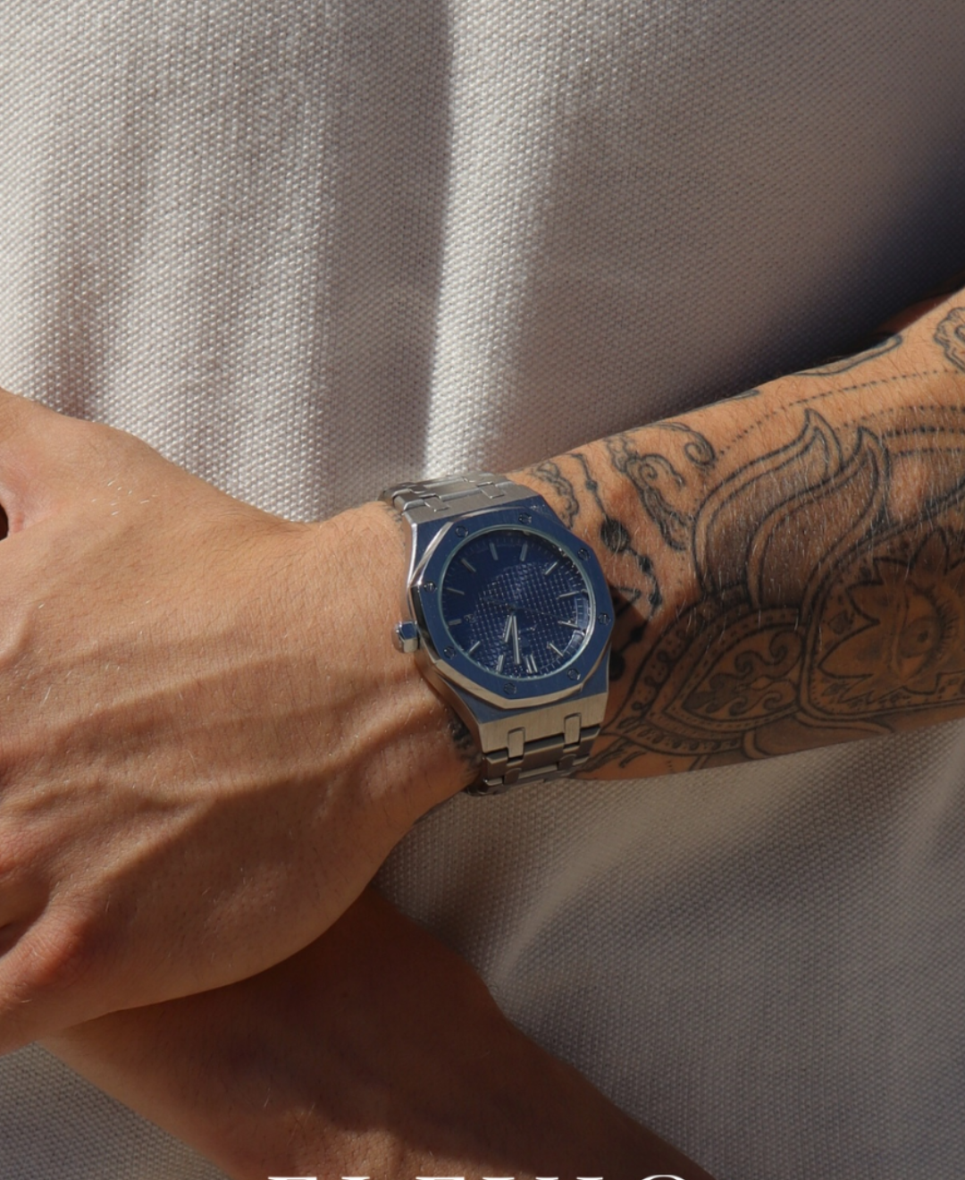 Close-up of a person's arm with tattoos wearing a silver-tone watch featuring a blue textured dial and a matching metal bracelet, against a light-colored polo shirt.