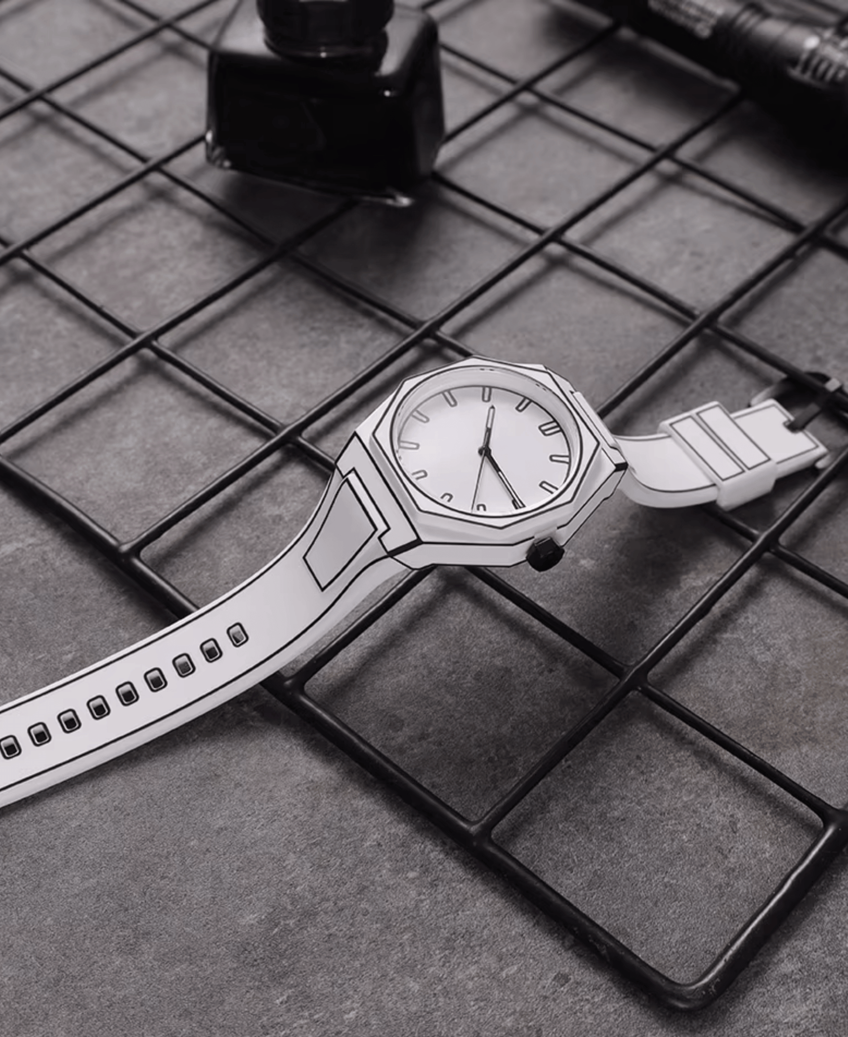 A white watch with black outline details, resembling a sketch, lying on a dark grey metal grid surface. The watch has a white face, black hour markers, and a white strap. An ink bottle and a pen are visible in the blurred background.