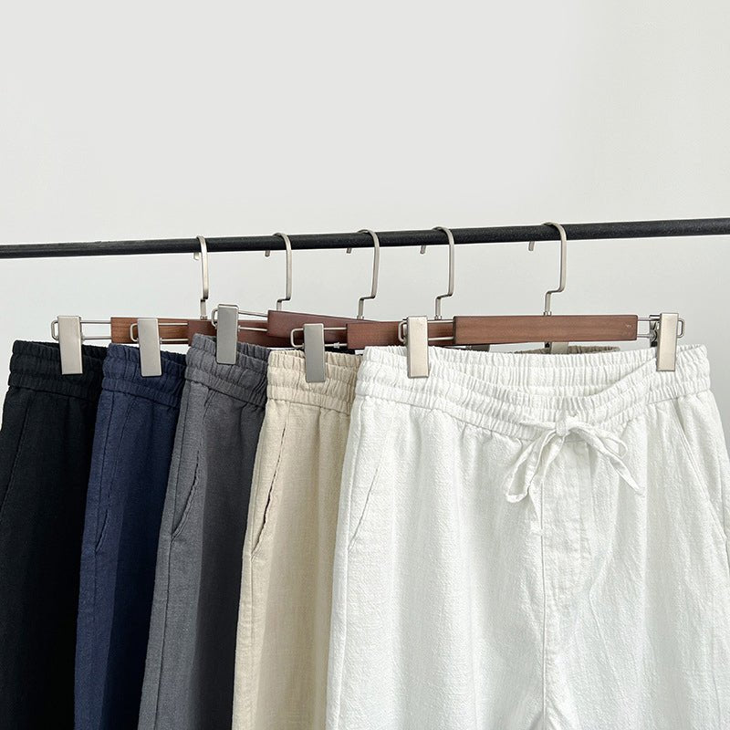 Close-up view of five pairs of Luxe Retreat Comfy Linen Pants with Relaxed Fit hanging on a rack, highlighting the elastic waistbands, drawstring, and side pockets in black, navy blue, gray, khaki, and white.