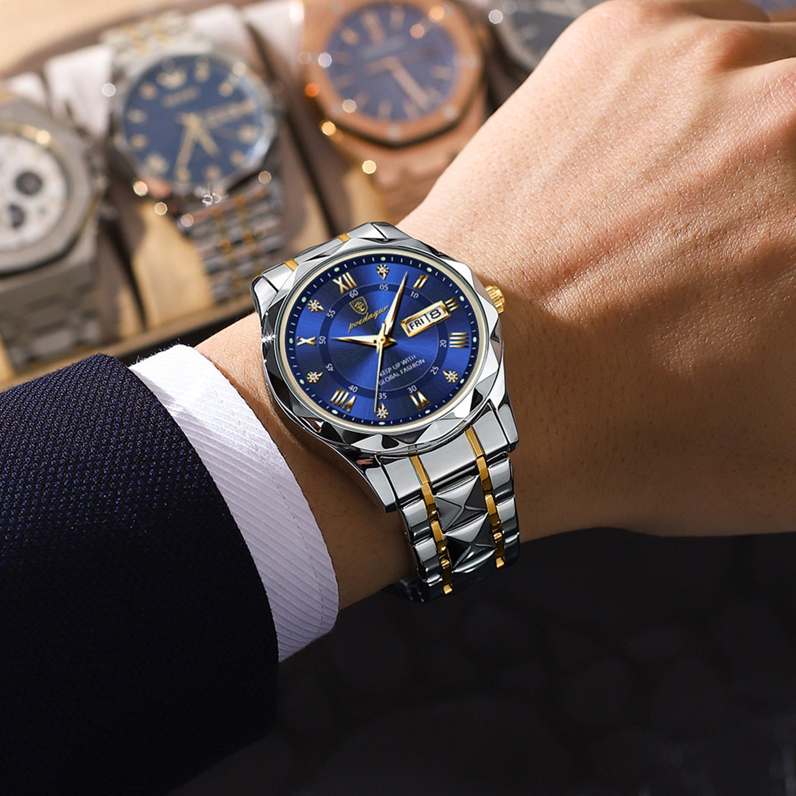 Maverick Luxe men's watch in stainless steel and gold, featuring a blue dial with date display and luminous hands, worn on a man's wrist with a suit jacket. Other watches are blurred in the background.