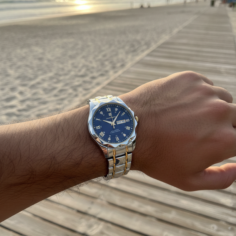 Maverick Luxe men's watch in stainless steel gold two-tone with blue dial and date display, worn on a man's wrist on a wooden boardwalk at sunset.