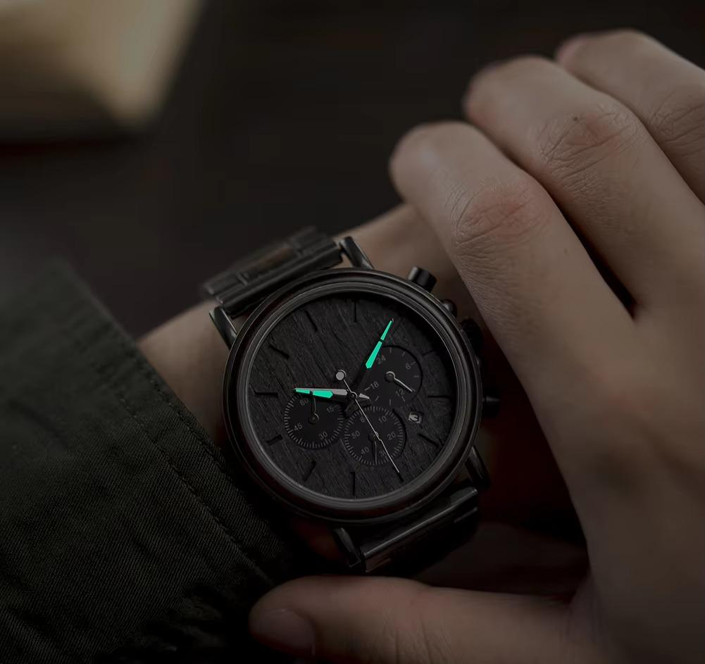 A man's wrist wearing the Nero Collection men's wooden watch, showcasing its minimalist design and the luminous green glow of the watch hands in a dimly lit environment.