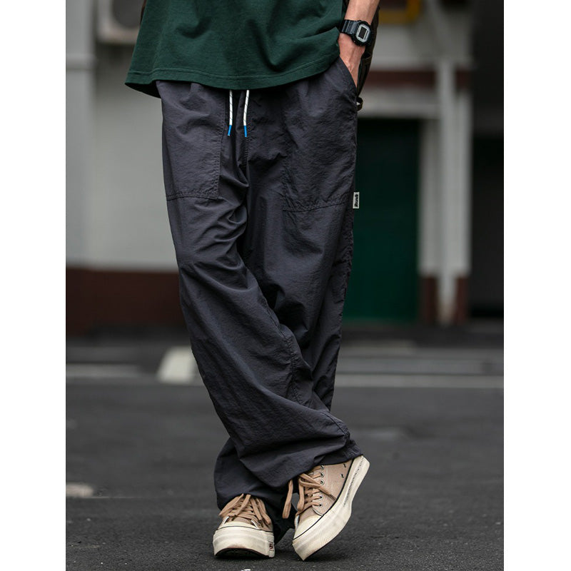 A man models Sakura Breeze waterproof, breathable, relaxed fit casual pants in deep grey, paired with a green t-shirt and canvas shoes, standing outdoors.