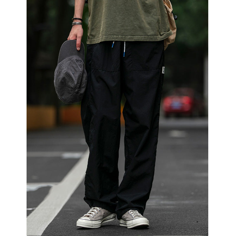 A man wearing black Sakura Breeze waterproof breathable casual pants with a relaxed fit, a green t-shirt, and sneakers, holding a cap. The pants are shown in a casual outdoor setting.