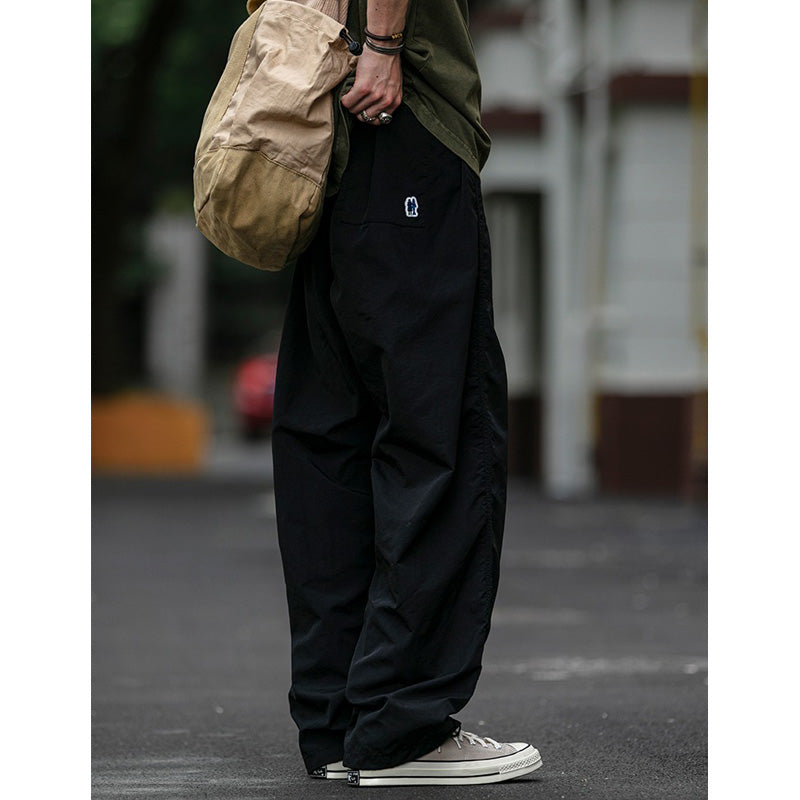 Side view of a man wearing black Sakura Breeze waterproof breathable casual pants with a relaxed fit, a green t-shirt, sneakers, and a beige shoulder bag. A small white logo is visible near the back pocket.