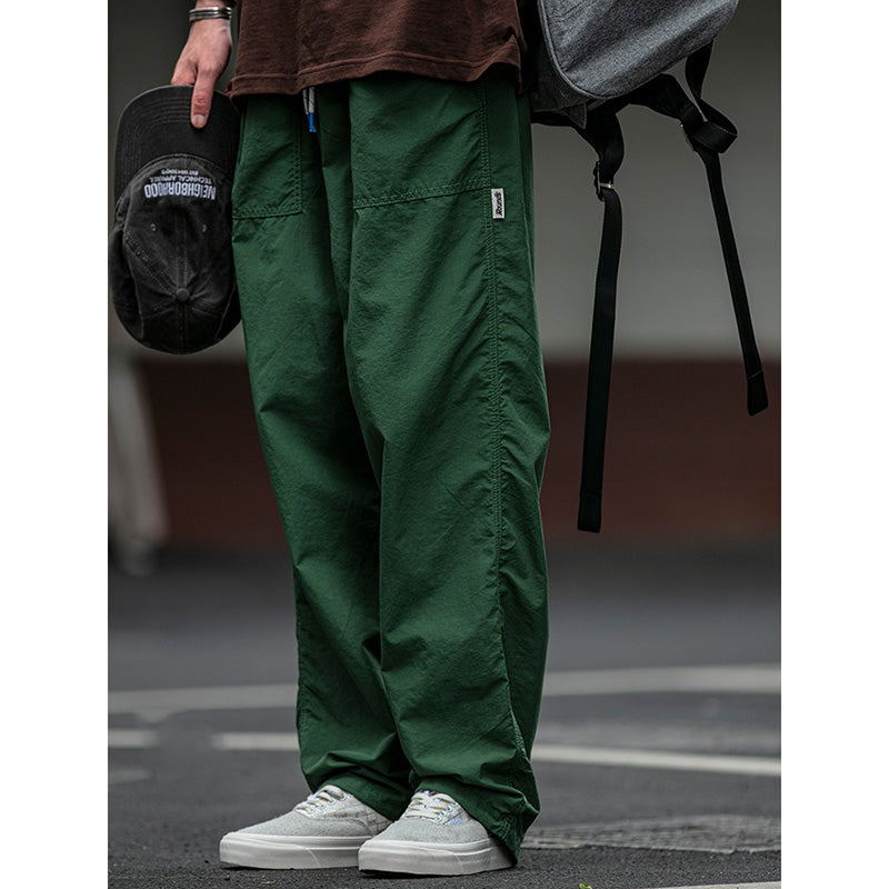 Side view of men's Sakura Breeze waterproof breathable casual pants in green, worn by a model with white sneakers and a brown t-shirt, holding a black cap. Showcases the relaxed fit and fabric texture.