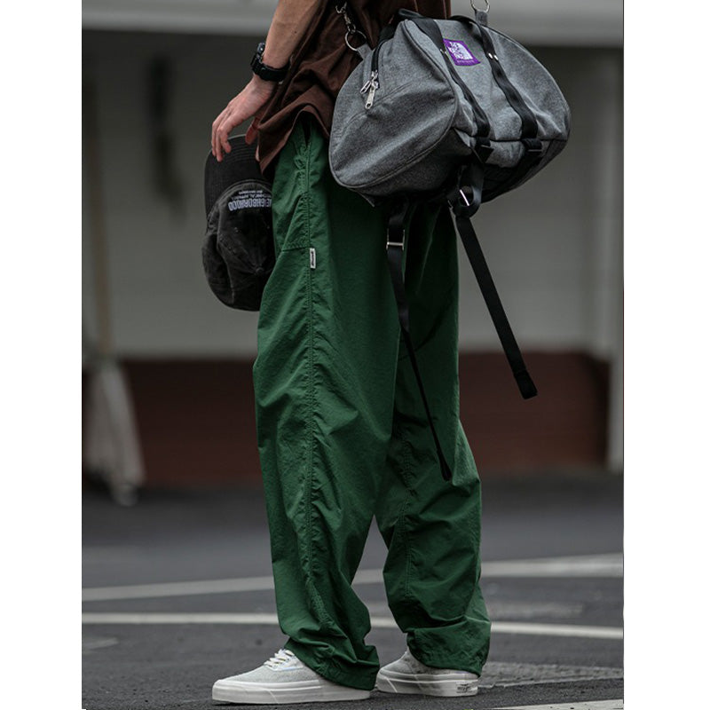 Side profile of men's Sakura Breeze waterproof breathable casual pants in green, worn by a model carrying a grey duffel bag and holding a black cap. Highlights the relaxed fit and overall casual outfit.