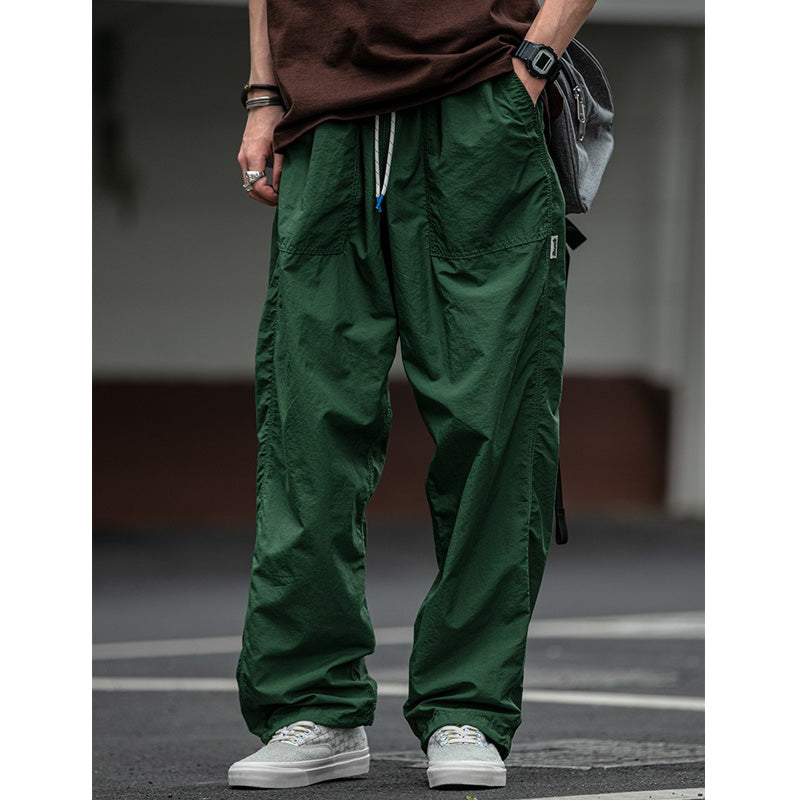 Men's Sakura Breeze waterproof breathable casual pants in green, shown on a model wearing white sneakers and a brown t-shirt, standing outdoors. Highlights the relaxed fit and drawstring waist.