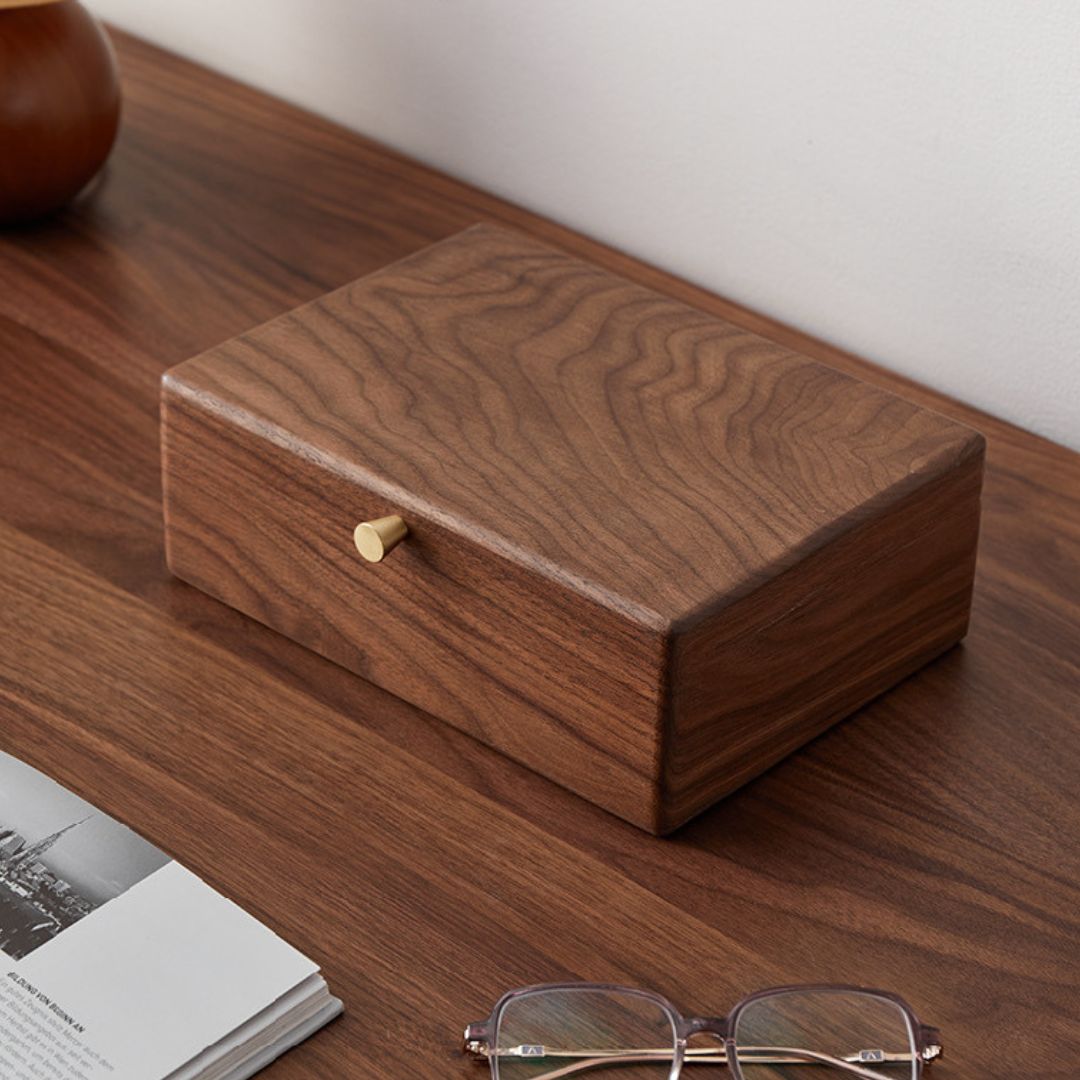 Closed solid walnut wood jewelry box with a brass knob, resting on a dark wooden desk alongside an open book and eyeglasses, highlighting its elegant design and home decor appeal.