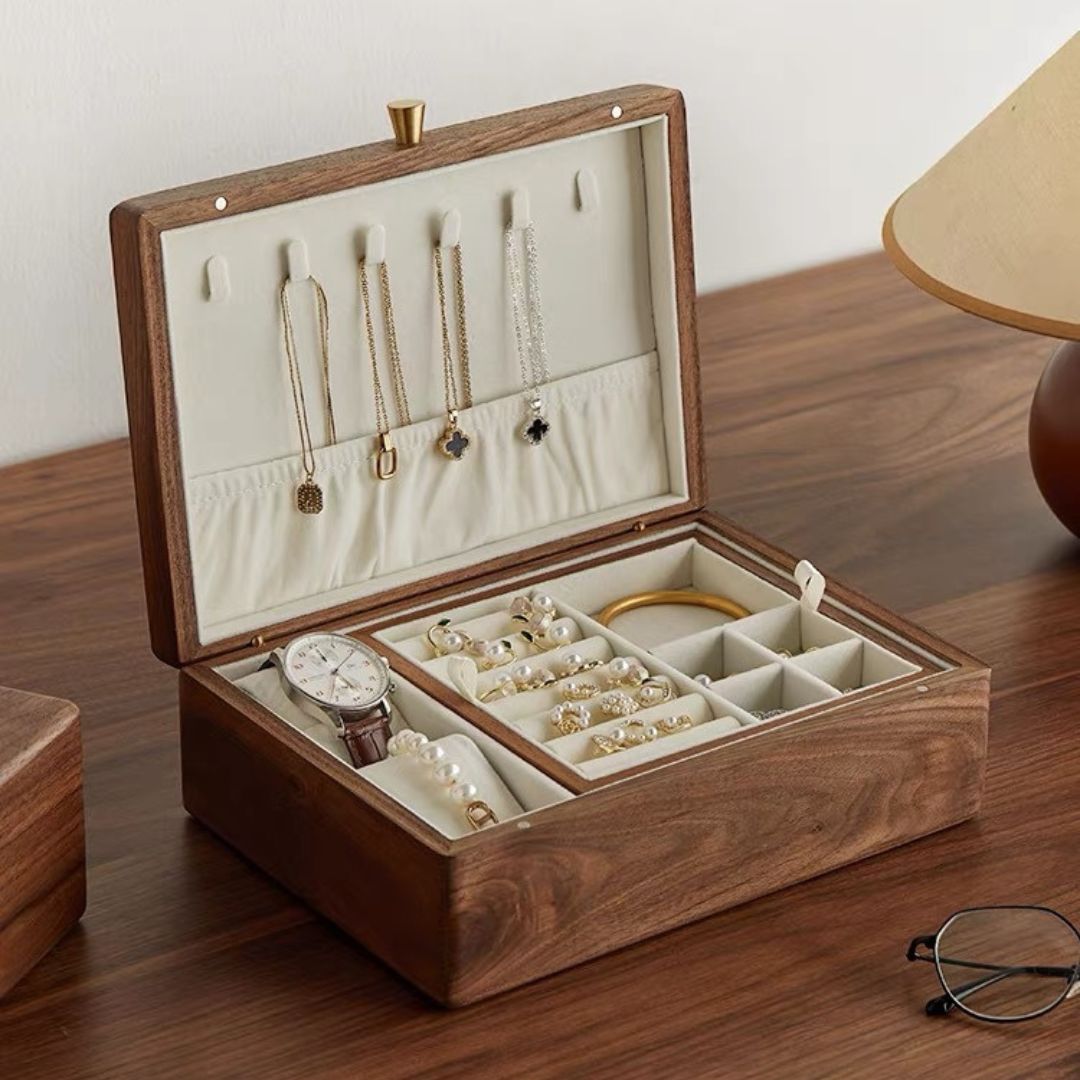 An open Timber Haven solid walnut wood jewelry box with a removable tray, filled with various jewelry items, placed on a wooden desk next to a lamp and reading glasses.