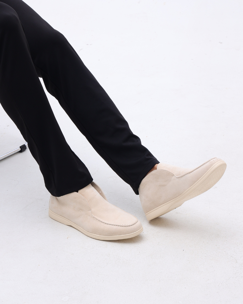 Beige suede loafers with classic detailing worn by a person in black pants, side view.