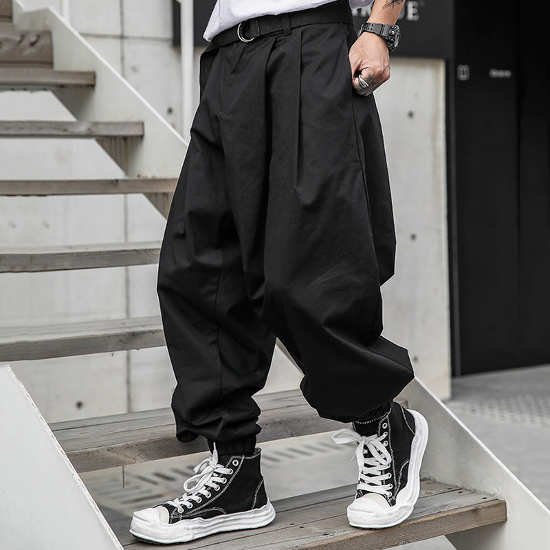 A man models Urban Essence men's pants in cool black with a retro design. The baggy, pleated pants feature elastic cuffs and are paired with a white t-shirt and black high-top sneakers, showcasing a comfortable, urban streetwear style on wooden stairs.