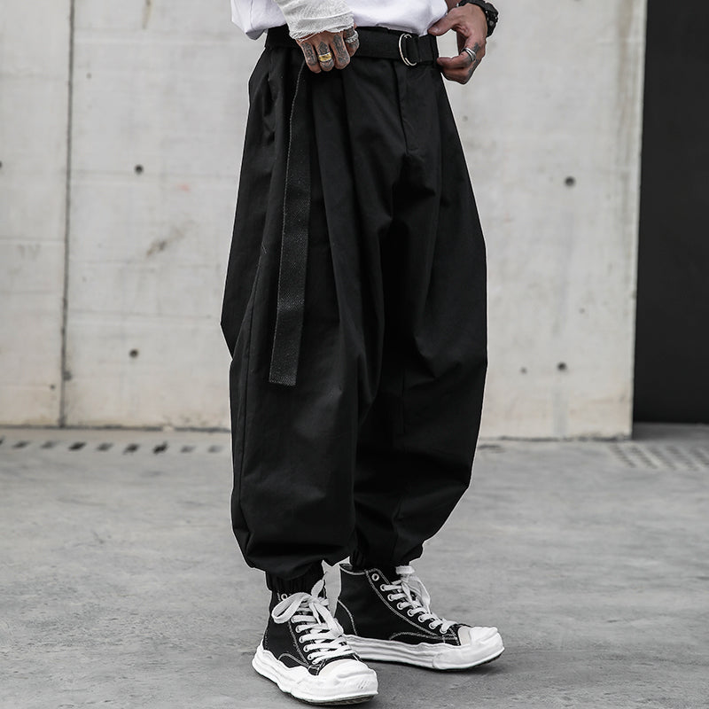 Side view of a man modeling Urban Essence men's pants in cool black with a retro design. The baggy, pleated pants feature elastic cuffs and a long belt, paired with a white t-shirt and black high-top sneakers, set against a concrete wall, highlighting the urban streetwear aesthetic.