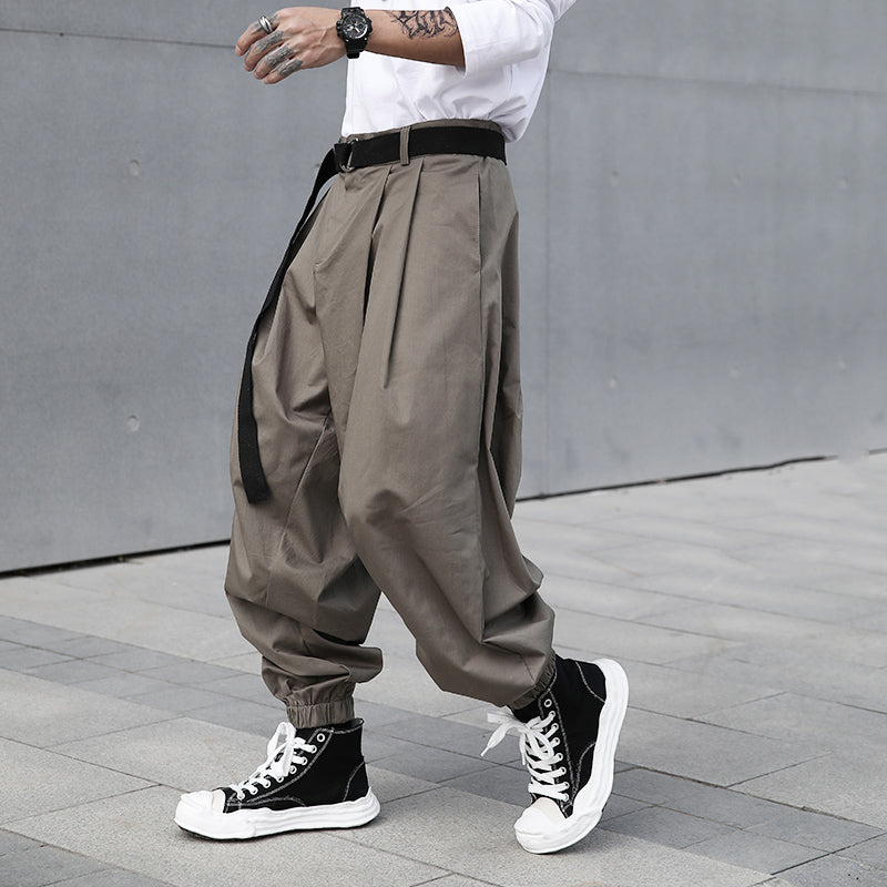 Male model walking, showcasing Urban Essence men's grey-green baggy pleated pants with a retro design, elastic cuffs, and a black belt, viewed from the side.