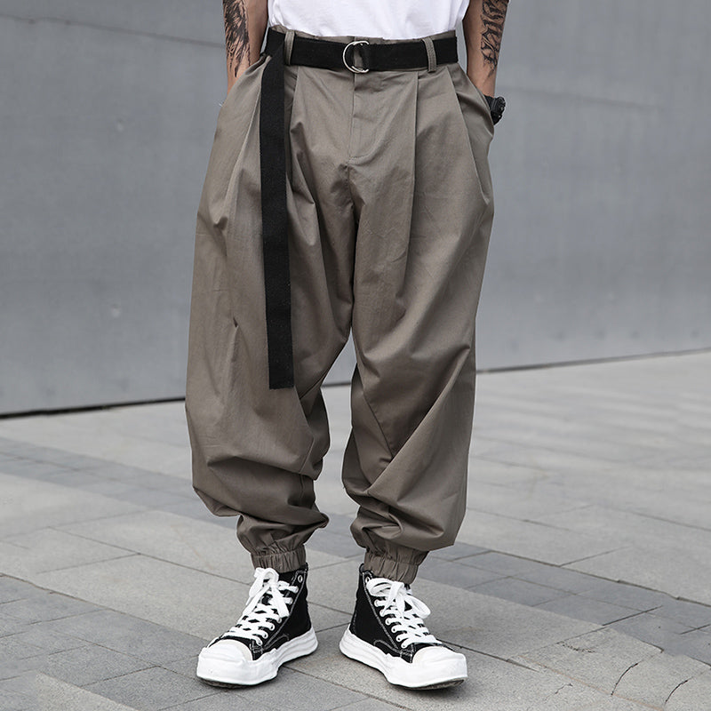 Front view of a male model wearing Urban Essence men's grey-green baggy pleated pants with a retro design, elastic cuffs, and a black belt.
