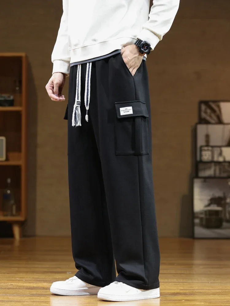 A person wearing black Urban Nomad cargo sweatpants made of soft cotton, featuring a large utility pocket on the left thigh and visible drawstrings at the waist. The model is standing in a side profile, with hands in pockets, showcasing the relaxed fit and casual style of the pants. White sneakers are visible.