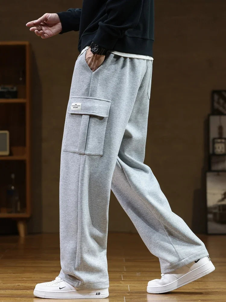 A person wearing light gray Urban Nomad cargo sweatpants made of soft cotton, featuring a large utility pocket on the left thigh. The model is standing in a side profile, with hands in pockets, showcasing the relaxed fit and casual style of the pants. White sneakers are visible.