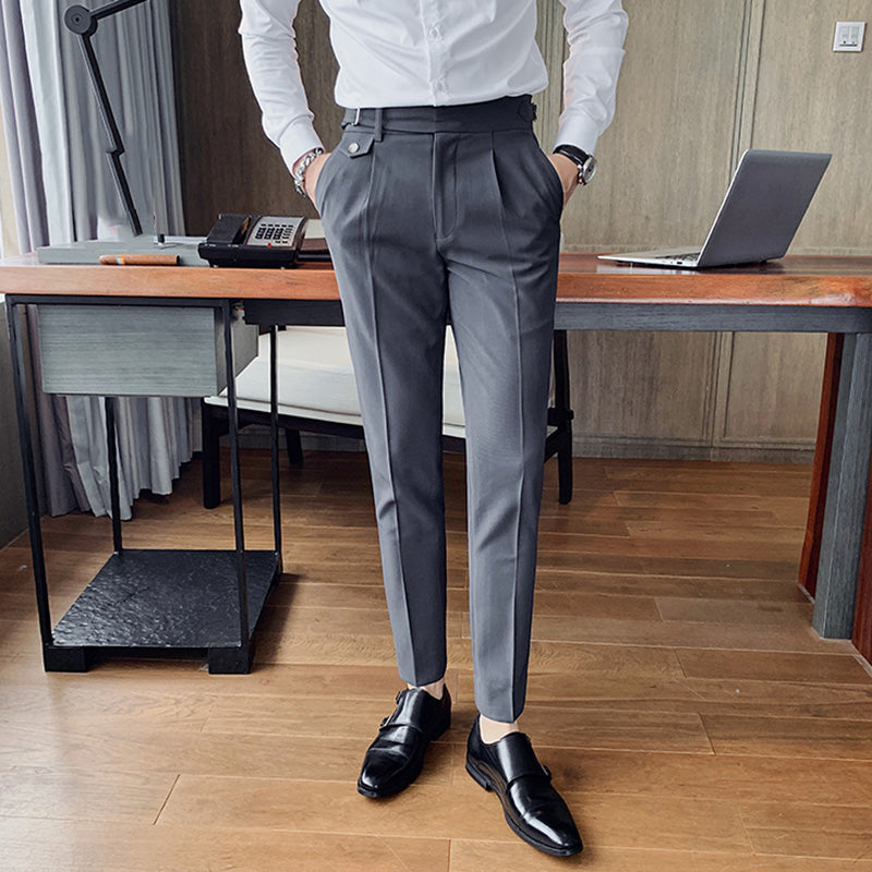 A man models Vintage Charm men's retro grey suit pants with a tailored fit and lightweight fabric, paired with a white shirt and black loafers in an office setting. The pants feature a unique side buckle detail.