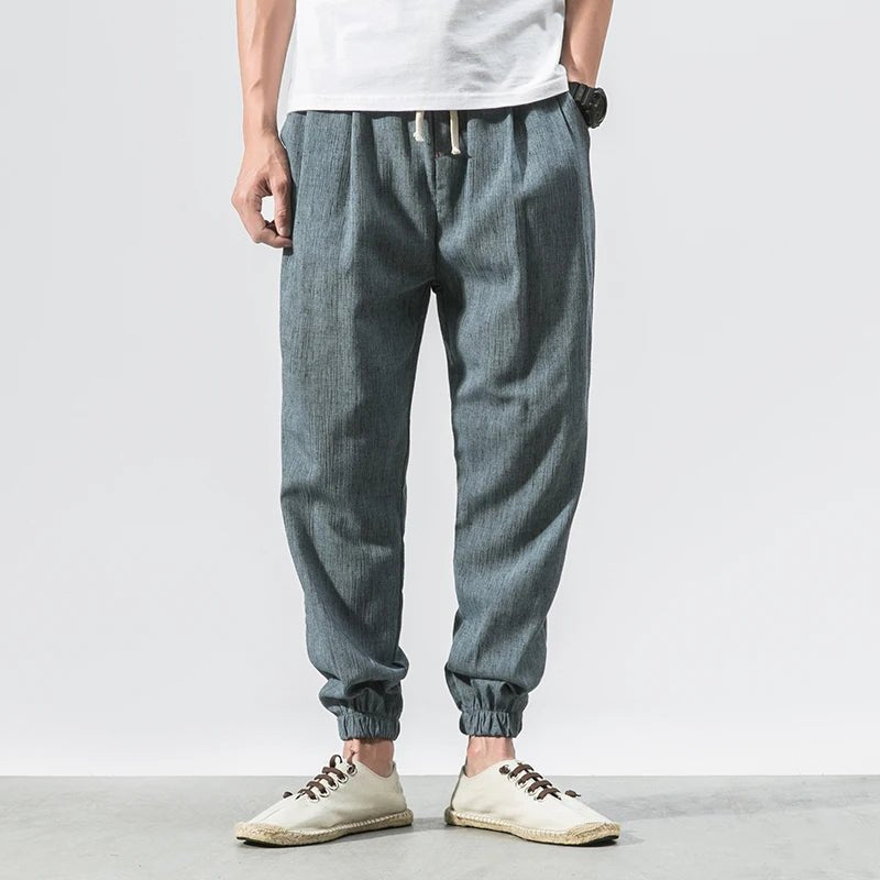 Full shot of a man wearing blue Wanderer's Path boho style linen pants with a relaxed fit, paired with a white t-shirt and light-colored slip-on shoes. The pants feature a drawstring waist and elastic cuffs, highlighting their comfortable and versatile design.
