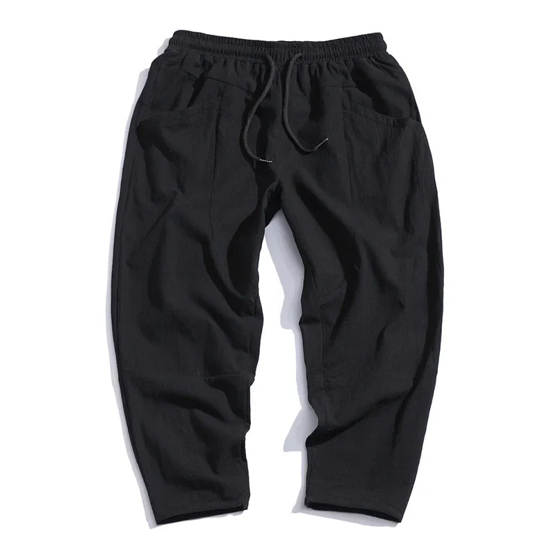 A pair of Zenith Cropped Trousers in black, made from a cotton-linen blend, laid flat. The image highlights the elastic drawstring waist and functional front pockets of the tailored, lightweight pants.