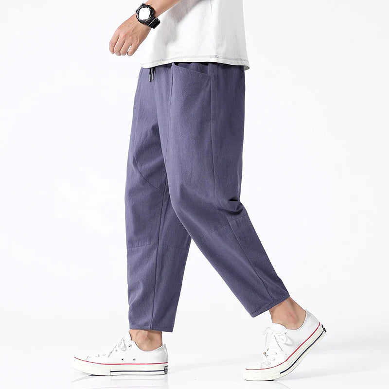 A man models Zenith Cropped Trousers in indigo, made from a cotton-linen blend, paired with white sneakers. The lightweight, tailored pants feature a drawstring waist and side pockets, perfect for smart-casual summer wear.