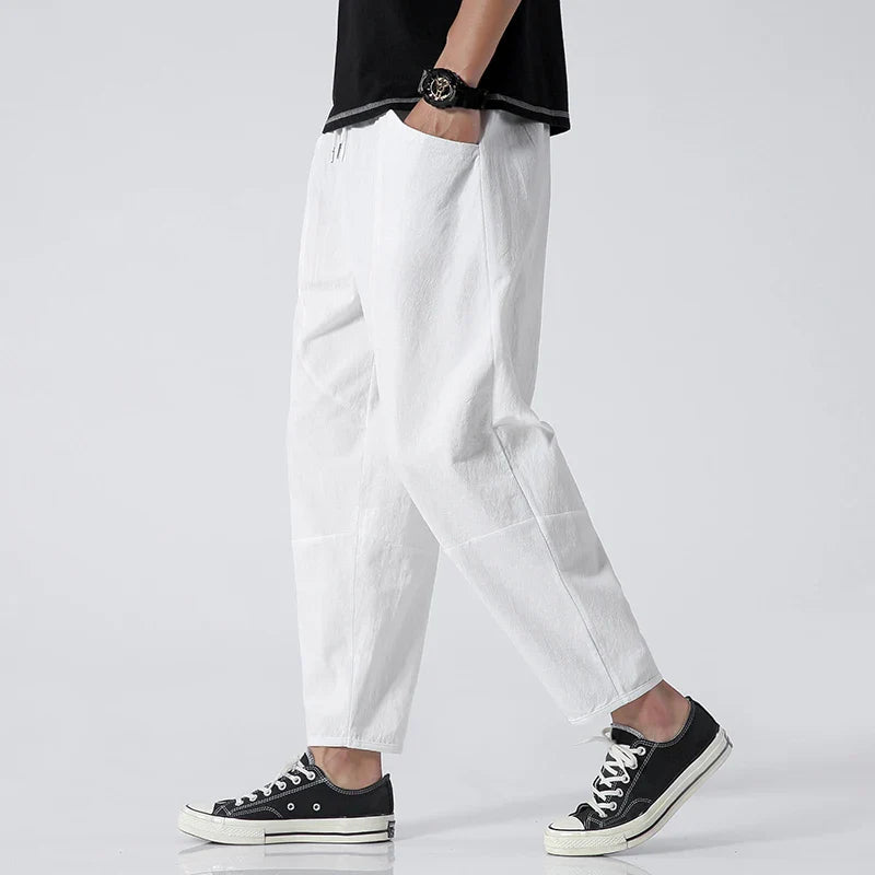 A man models Zenith Cropped Trousers in white, made from a cotton-linen blend, paired with black sneakers. The lightweight, tailored pants feature a drawstring waist and side pockets, suitable for smart-casual summer outfits.