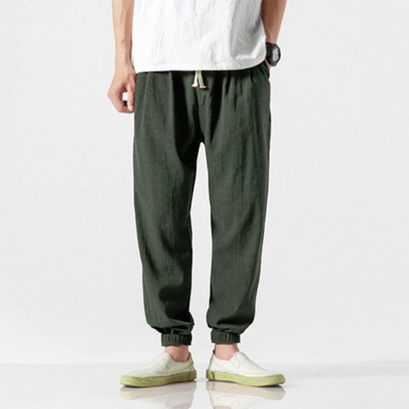 Full shot of a man wearing Zenith Stones dark olive green linen men's pants with a drawstring waist and elastic cuffs, styled with a white t-shirt and light green slip-on shoes.