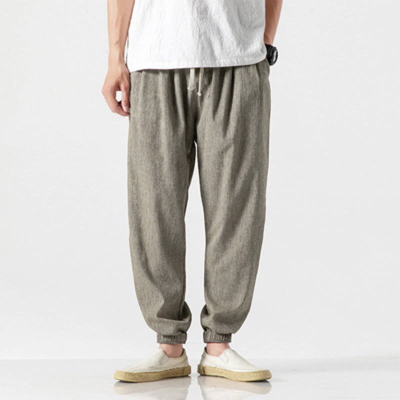 Full body shot of a man wearing Zenith Stones DarkKhaki linen drawstring pants with elastic cuffs, paired with a white t-shirt and light-colored casual shoes. The pants feature side pockets and a relaxed fit.
