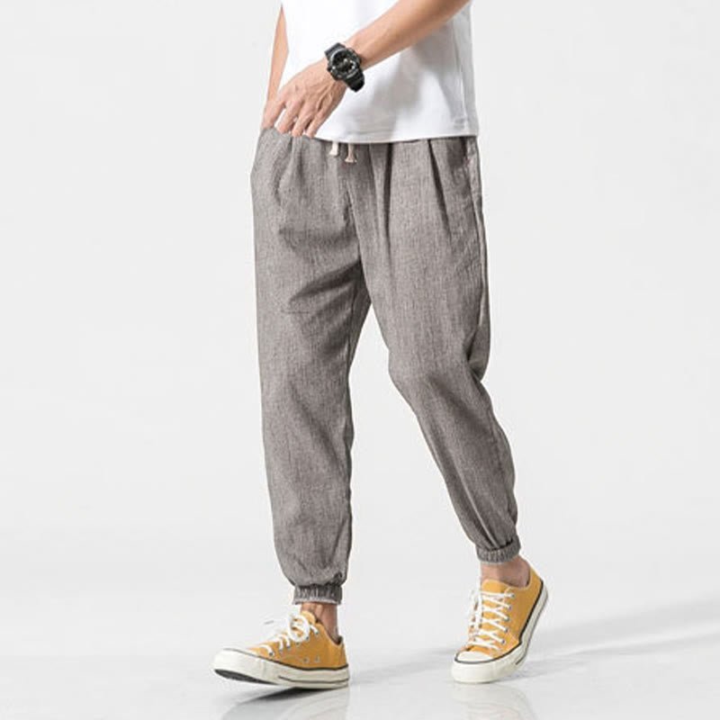 A man models Zenith Stones gray linen drawstring pants with side pockets and elasticated cuffs, paired with yellow sneakers. He stands with his hand on his hip, highlighting the comfortable, relaxed fit.