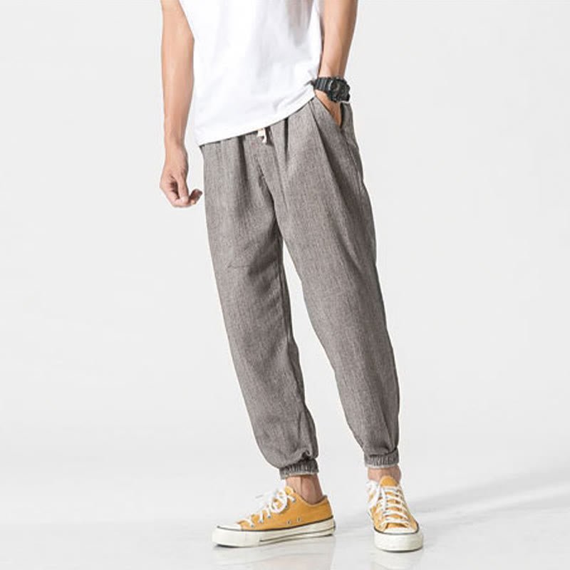 A man models Zenith Stones gray linen drawstring pants with side pockets and elasticated cuffs, paired with yellow sneakers. He stands with one hand in his pocket, showcasing the relaxed fit and casual style of the pants.