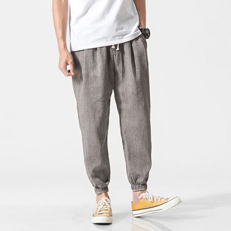 A man models Zenith Stones gray linen drawstring pants with side pockets and elasticated cuffs, paired with yellow sneakers. He stands facing forward with both hands on his hips, showcasing the casual style.