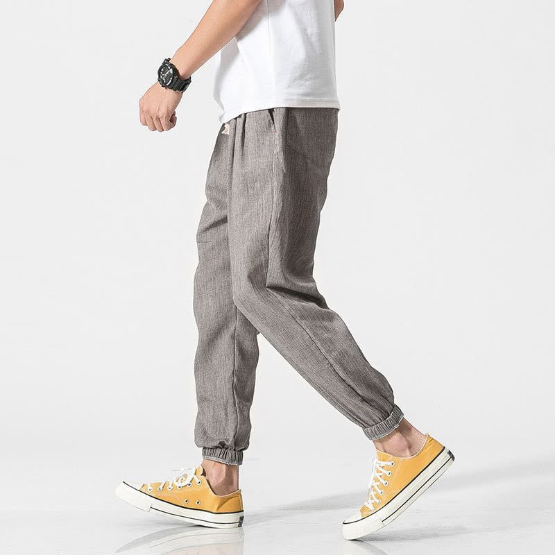 A man models Zenith Stones gray linen drawstring pants with side pockets and elasticated cuffs, paired with yellow sneakers. He is captured mid-stride, demonstrating the comfortable movement of the lightweight fabric.
